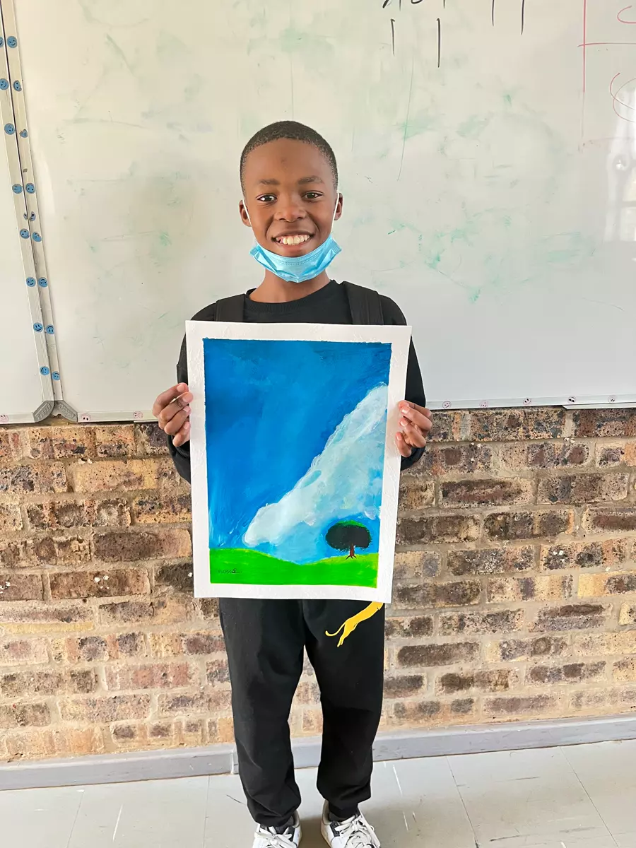 A young boy holding up his painting of a landscape with a blue sky and a tree