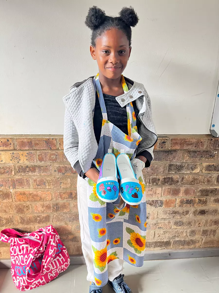 A young girl holding up painted shoes wearing an apron